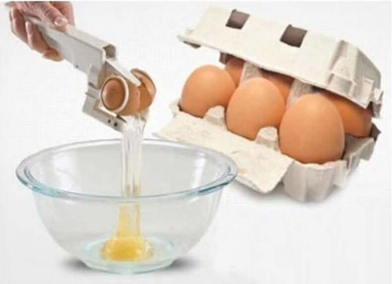 Handheld egg cracker tool breaking an egg into a bowl with a carton of eggs in the background.