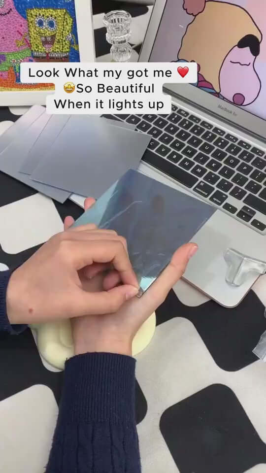 Person holding a shiny blue mirror sheet, with a laptop and colorful art in the background, showcasing DIY decor.